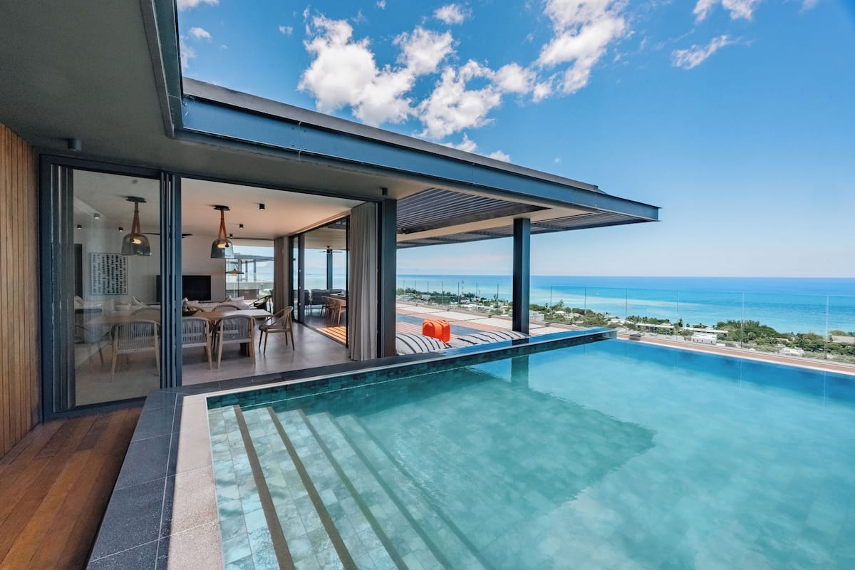 A modern poolside area with a clear view of the ocean, a covered seating area, and a glass-enclosed space with furniture.