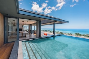 A modern poolside area with a clear view of the ocean, a covered seating area, and a glass-enclosed space with furniture. - Legend Hill Residences & Spa (Black River)