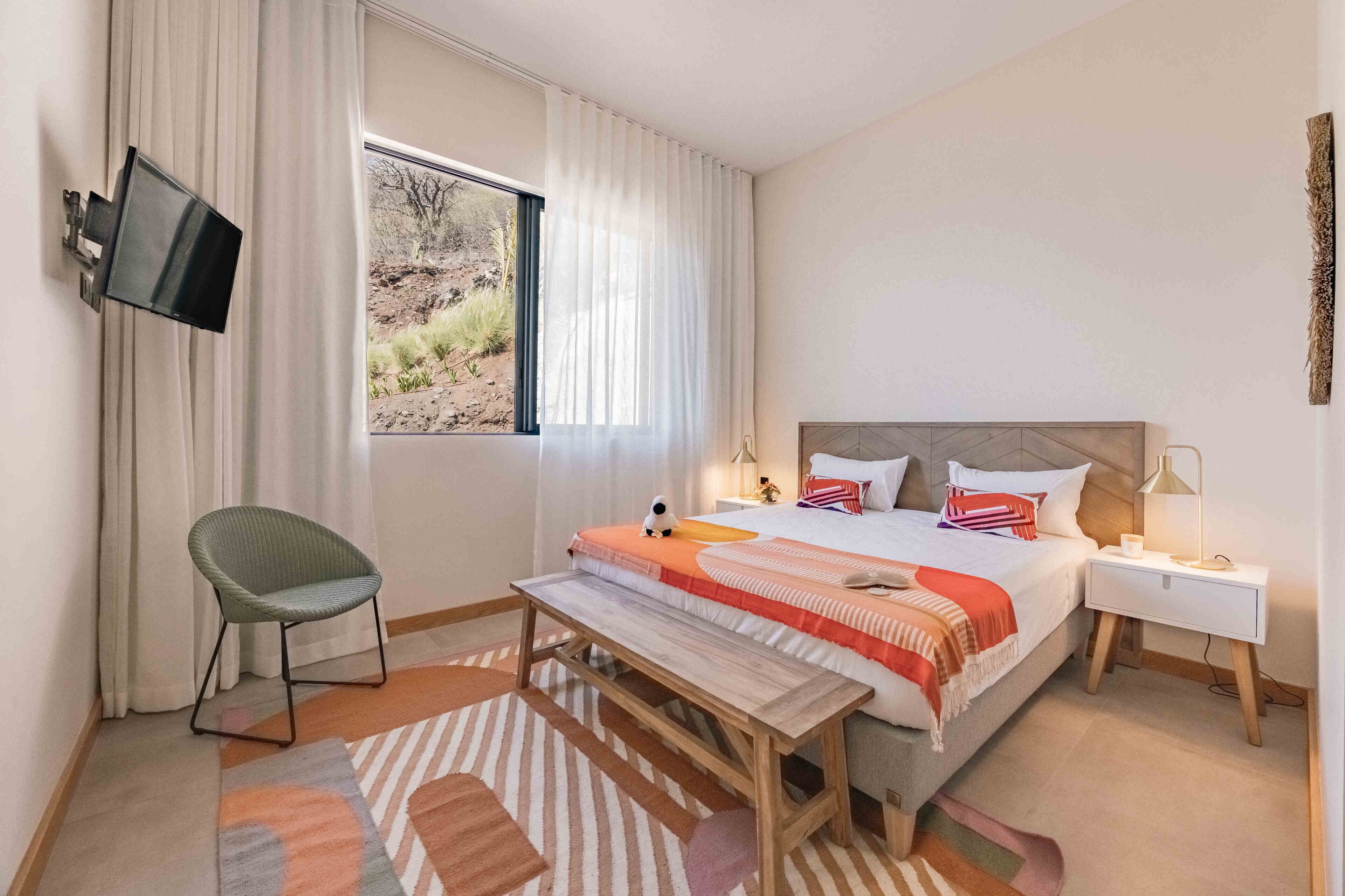 A modern hotel room with a large bed, a flat-screen TV mounted on the wall, a bedside table with a lamp, and a view of the outdoors through the window.