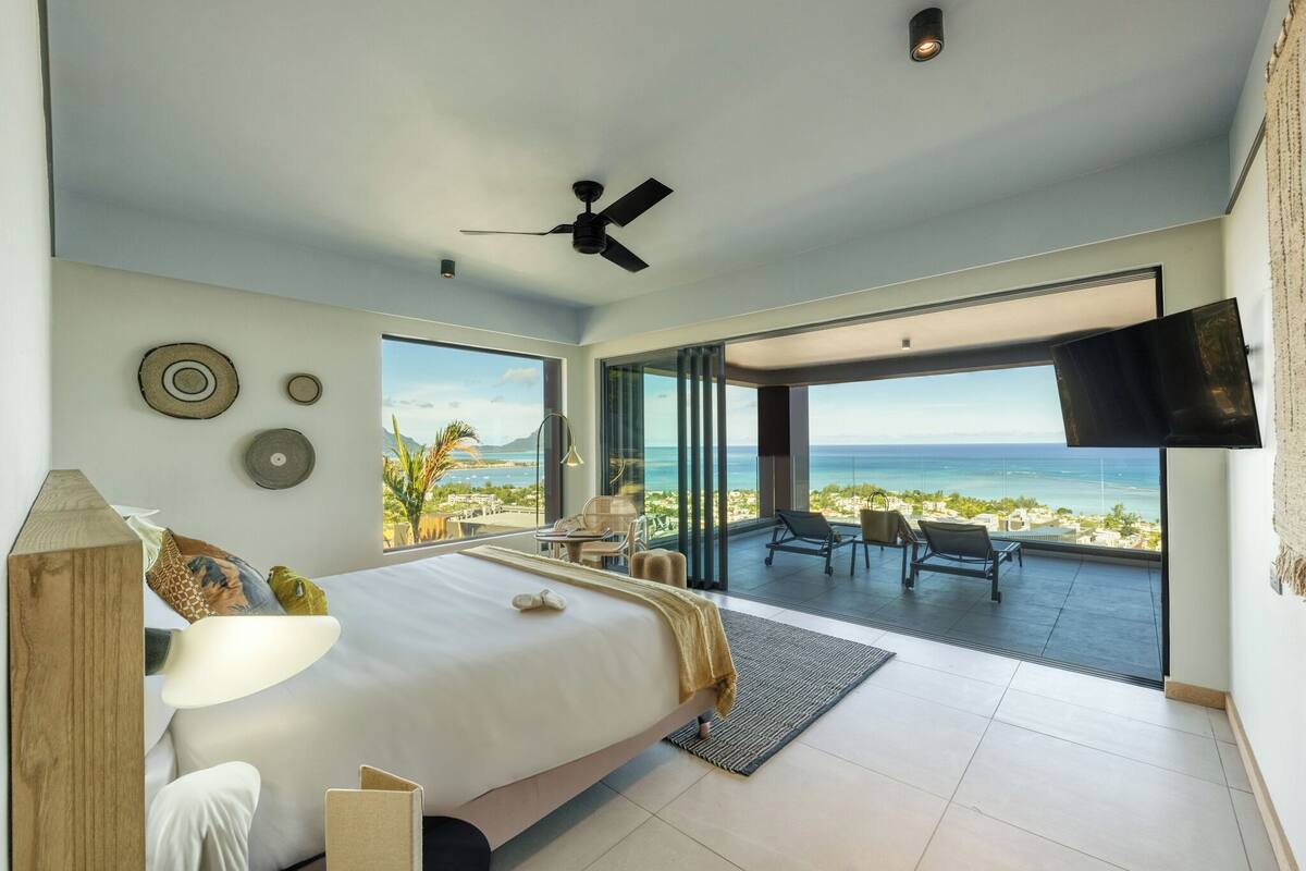 A modern bedroom with a large bed, a ceiling fan, and a view of the ocean.