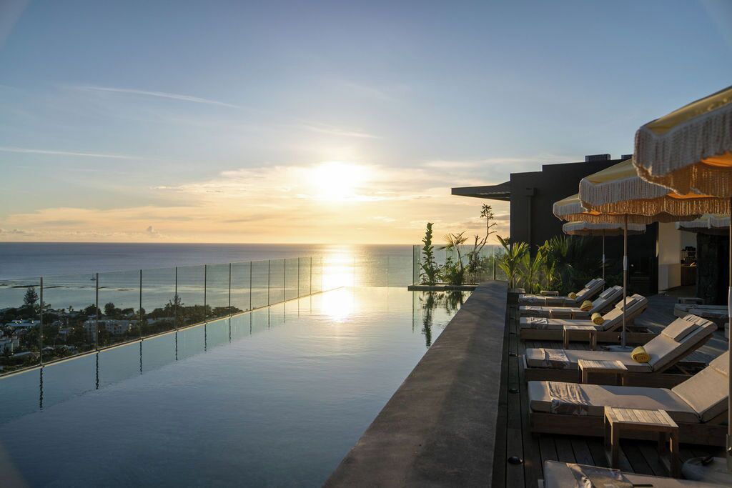 Una piscina infinita con sillones y sombrillas, con vistas a una puesta de sol sobre el océano.