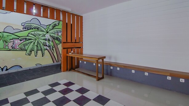 Ein Zimmer mit Schachbrettmuster-Boden, einer hölzernen Bank, einer Wand mit Palmen-Wandgemälde und einem Lüftungsgitter an der Decke.