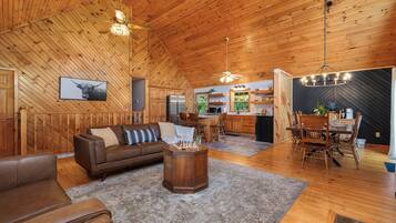 A spacious living room with a wooden ceiling, a brown leather sofa, a coffee table, and a dining area with wooden chairs and a chandelier.