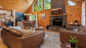 Amplia sala de estar con chimenea de piedra, techo de madera y ventanas grandes.