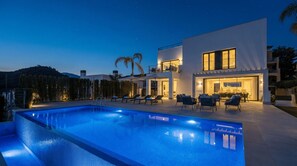 A modern poolside area with a swimming pool, lounge chairs, and a two-story building with outdoor lighting. - Villa de La Resina: A hidden gem and golfers paradise (Estepona)
