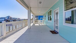 A covered outdoor area with a wooden deck, a white railing, and a view of a building with a blue facade in the background. - Laguna Cove Cabin, Canal Front (Bolivar)