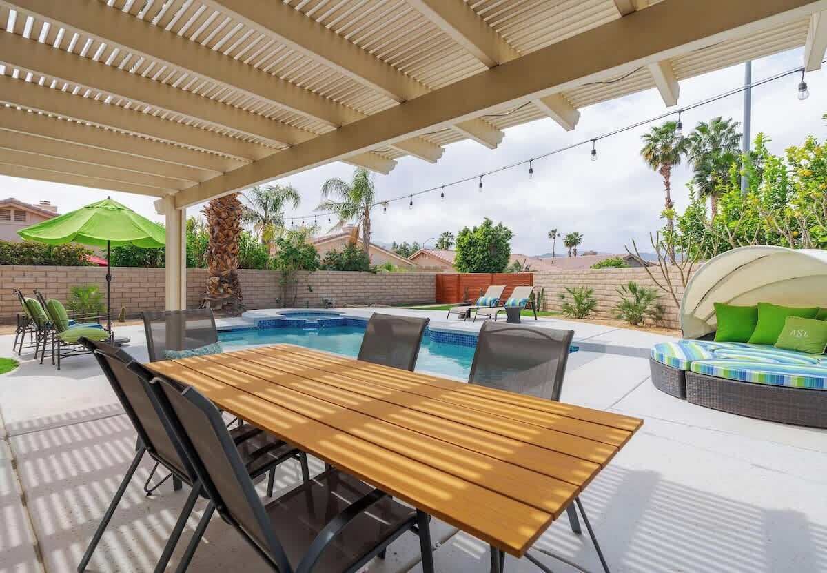 A covered outdoor area with a pool, wooden table, chairs, and a lounge area with cushions.