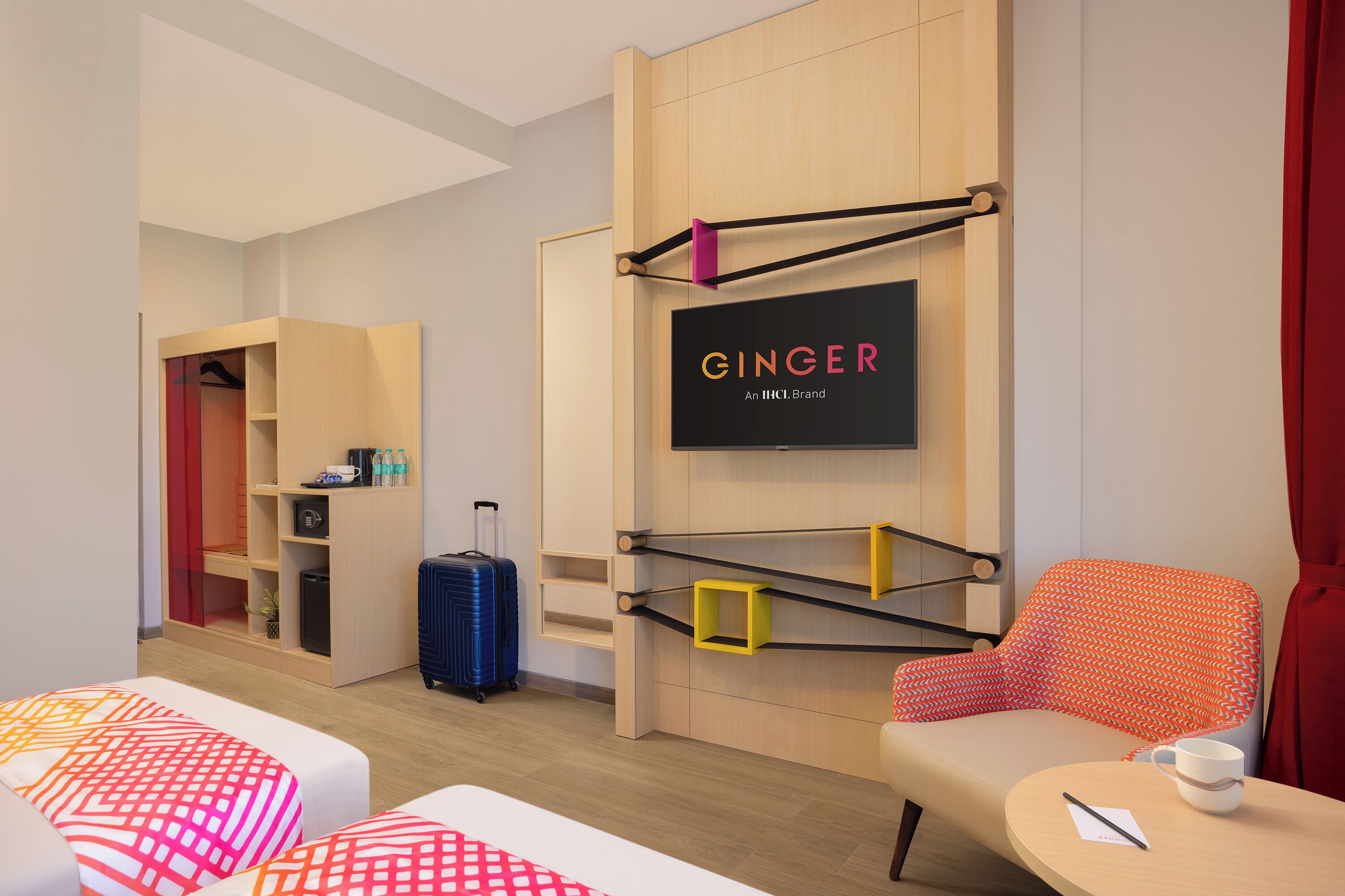 A hotel room with a bed, a chair, a small table, a suitcase, and a wall-mounted shelf with a sign.