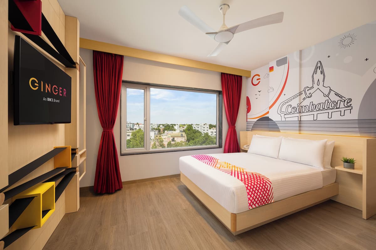 A modern hotel room with a large bed, a TV mounted on the wall, a window with red curtains, and a city view.