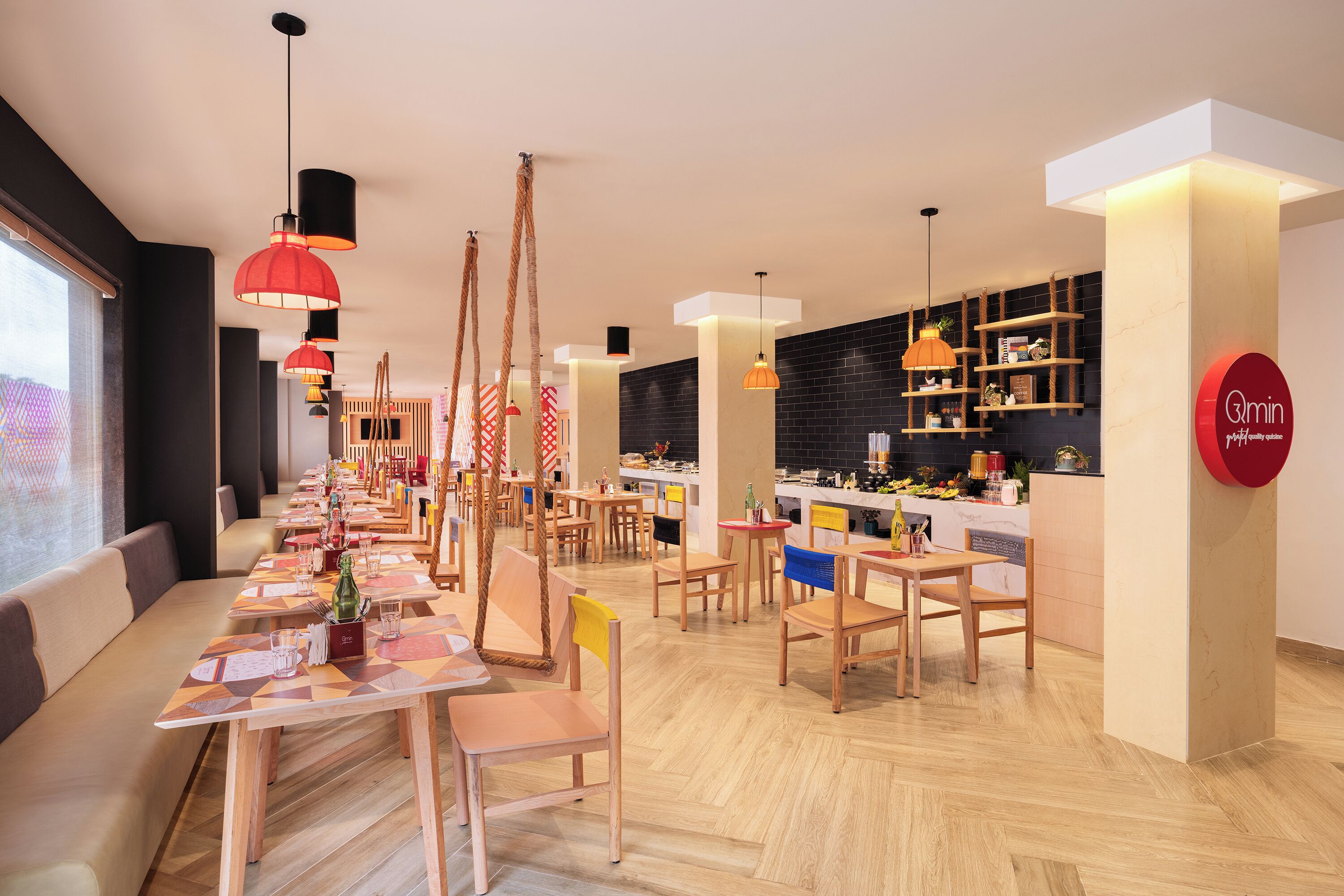 A modern restaurant with wooden tables and chairs, hanging red lamps, and a kitchen area with shelves.