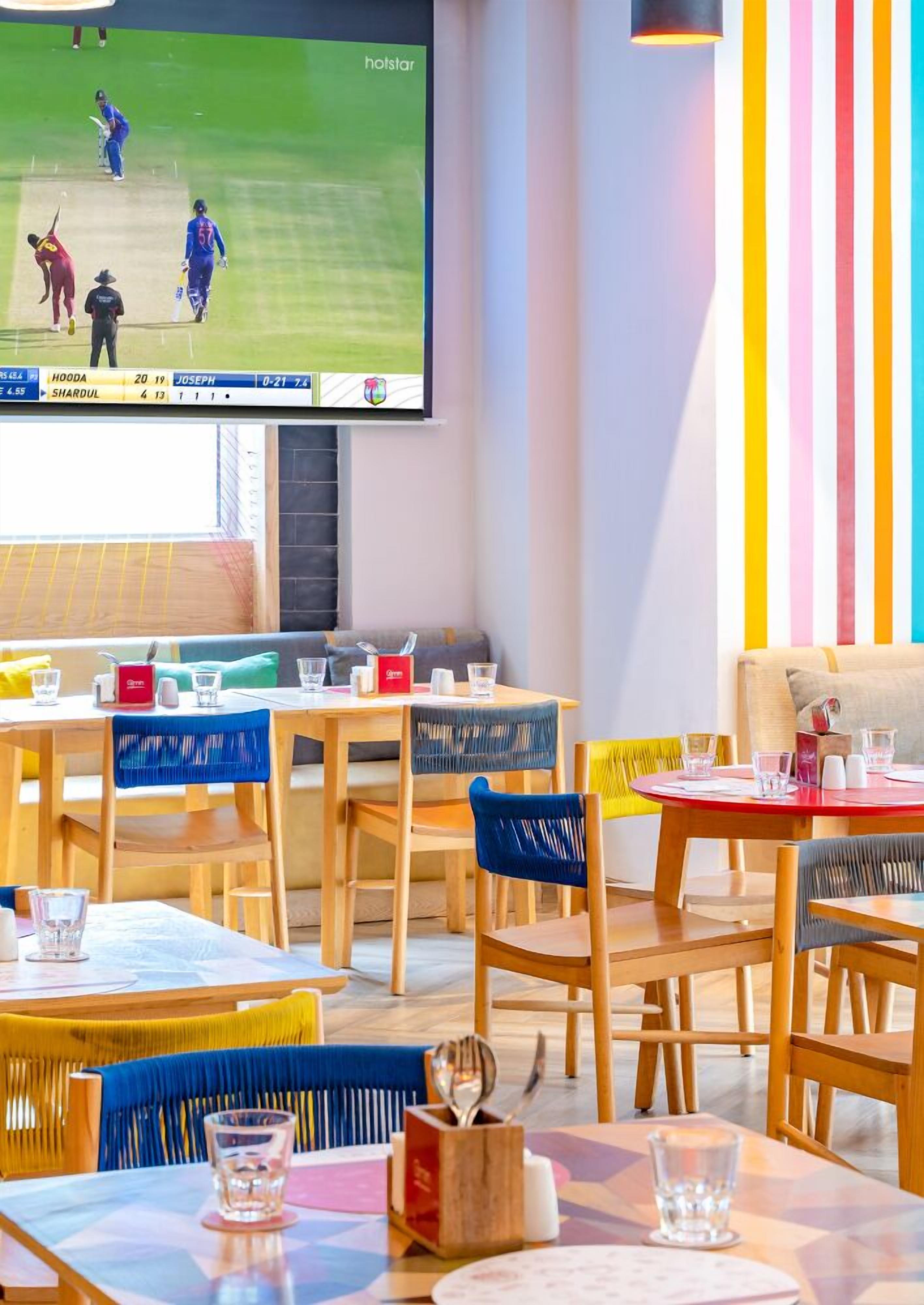A restaurant with wooden tables and chairs, a flat-screen TV displaying a cricket match, and a colorful striped wall.