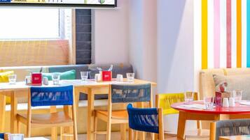 A restaurant with wooden tables and chairs, a flat-screen TV displaying a cricket match, and a colorful striped wall.