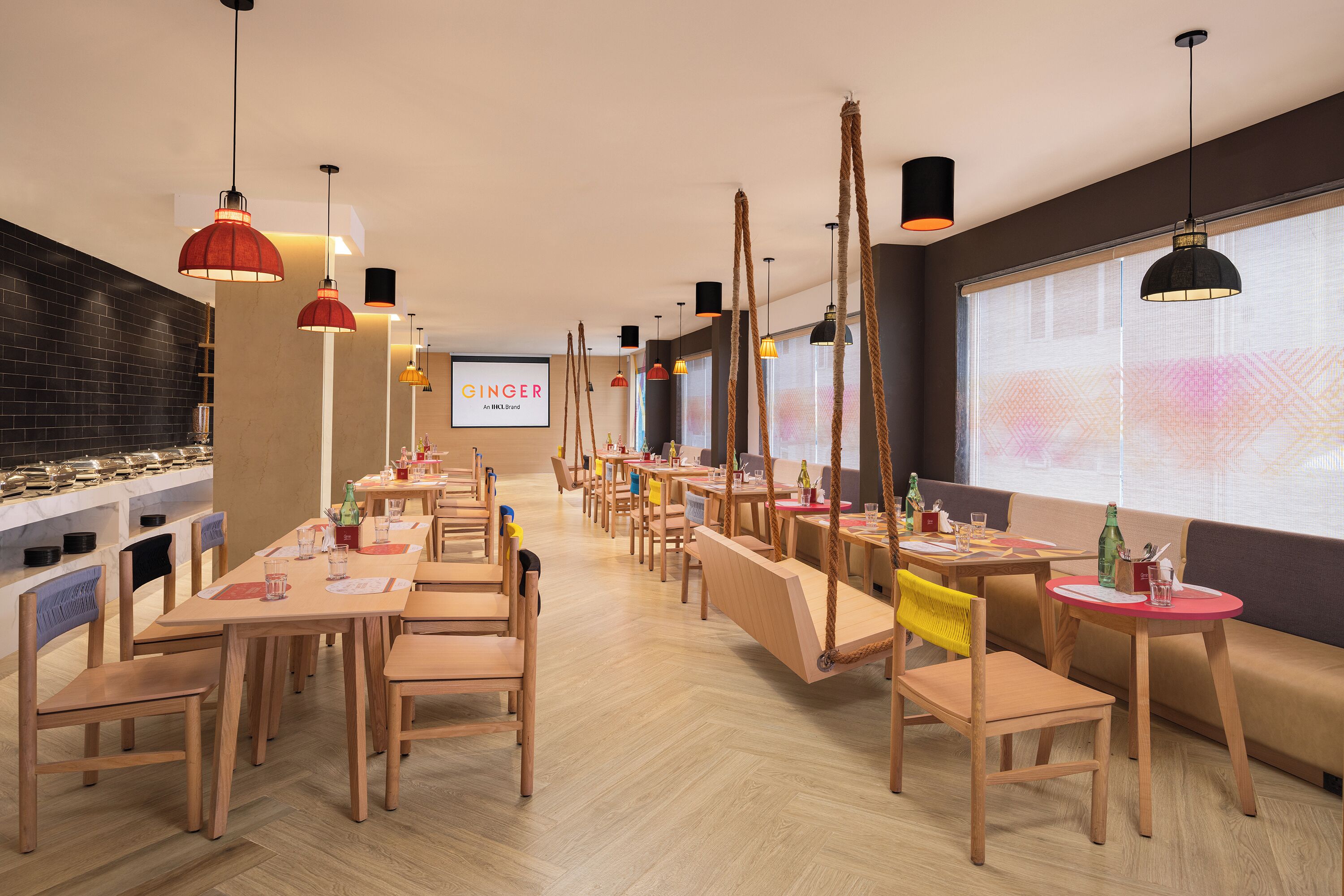A modern dining area with wooden tables and chairs, hanging pendant lights, and a large window with a view.