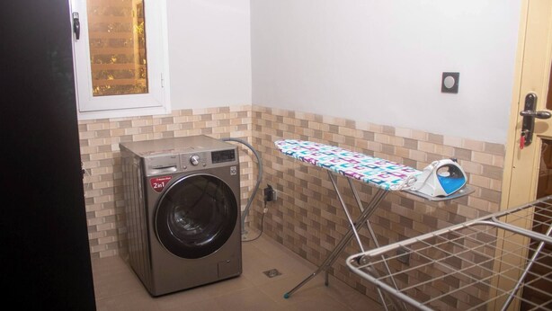 A compact laundry area with a washing machine, ironing board, and drying rack. - Kadoued Furnished Apartment 2 Bedroom (Ouagadougou)