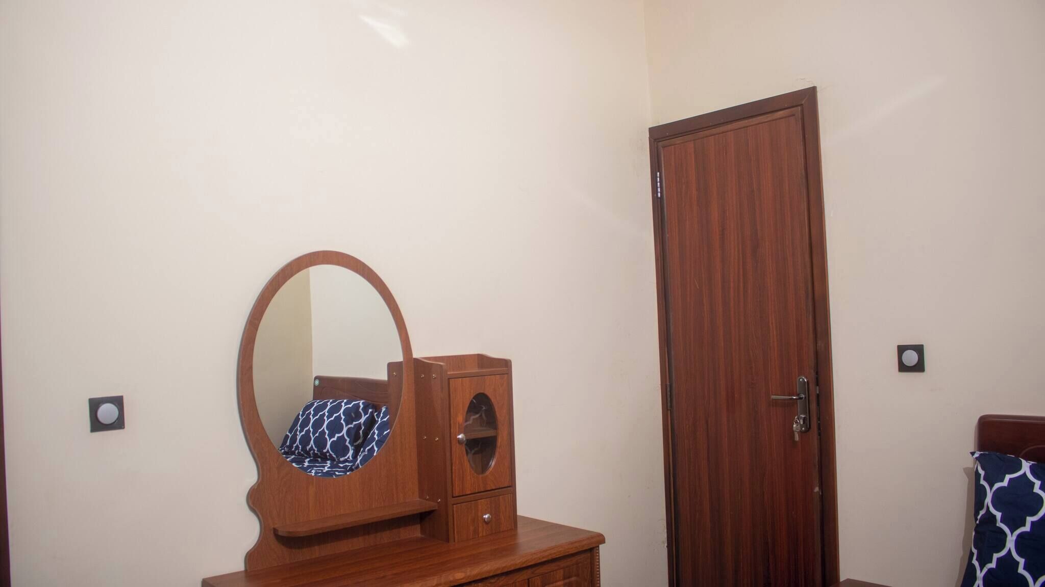 A room with a wooden dresser, a mirror, a door, and a patterned pillow.