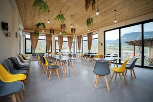 Un salón de conferencias con mesas y sillas, ventanas amplias y plantas colgantes.