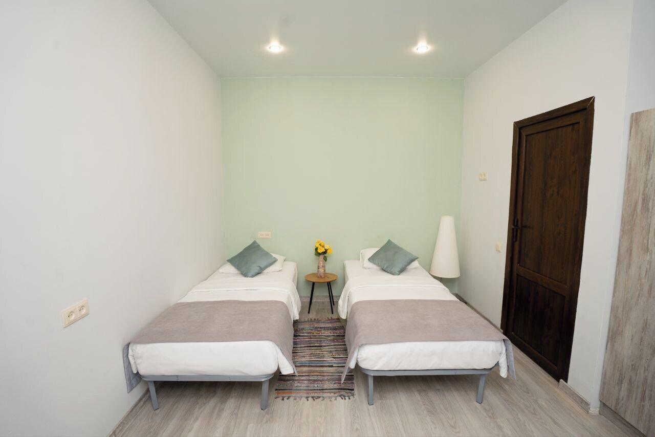 A small, well-lit room with two beds, a wooden door, and a small table with a vase of flowers.