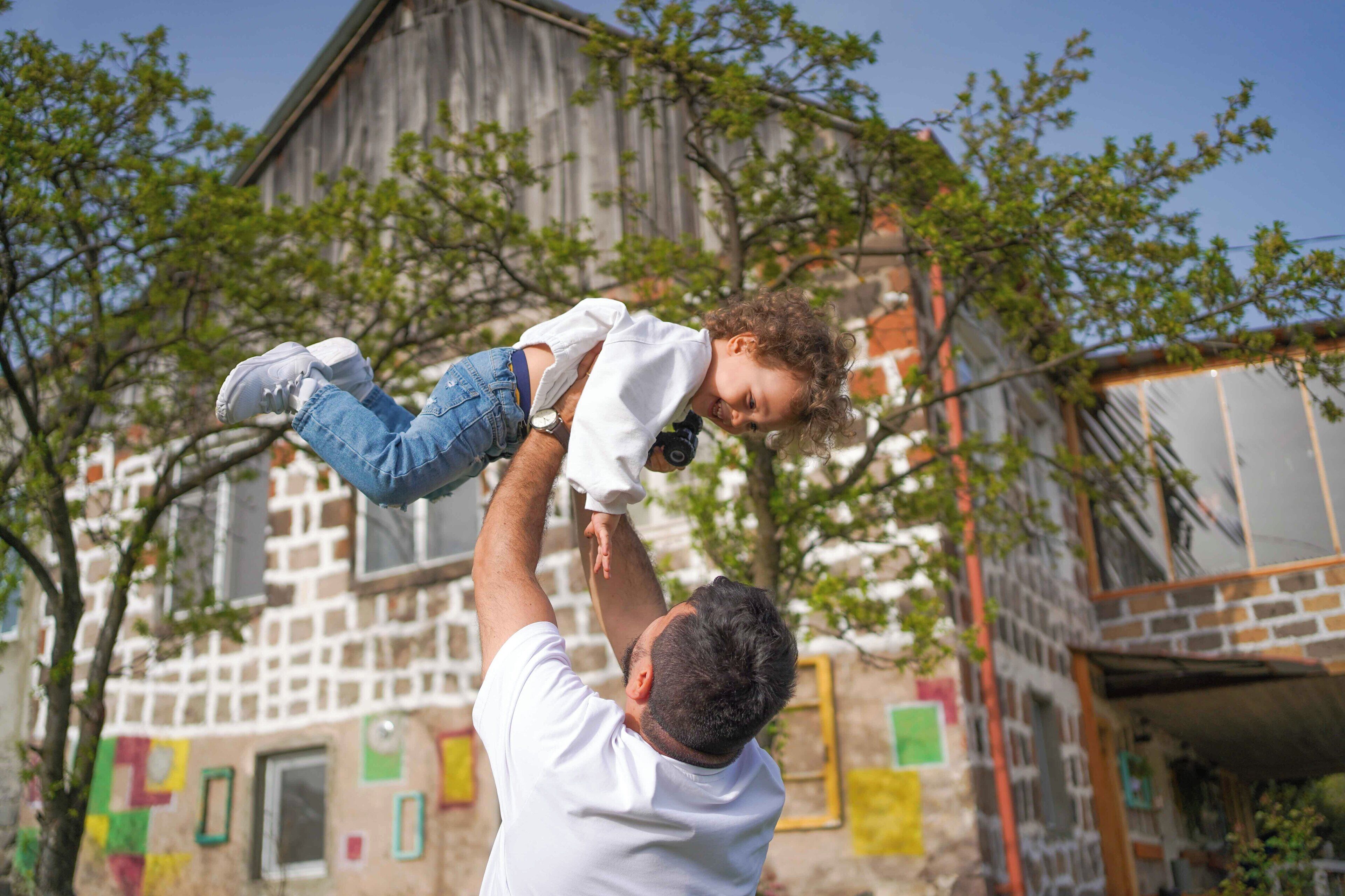 A person holding a child in an outdoor setting with a building in the background.