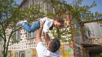 A person holding a child in an outdoor setting with a building in the background.
