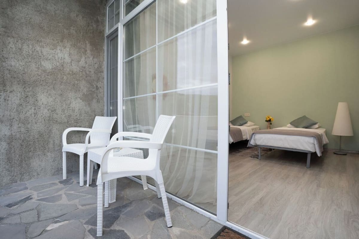 A room with a stone floor, two white chairs, a bed with pillows, and a lamp.