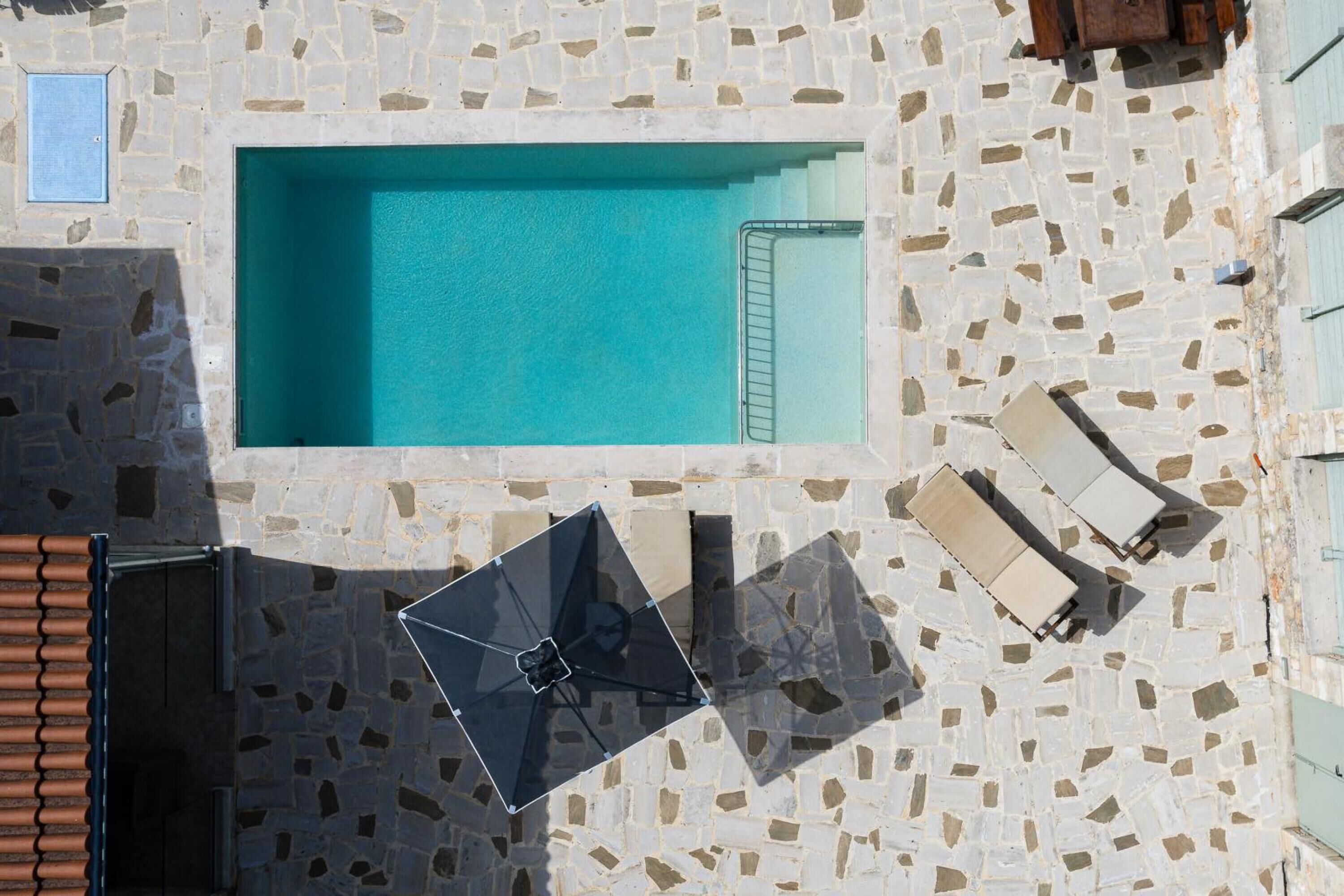 An aerial view of a pool area with a stone-tiled floor, a rectangular pool, and two lounge chairs.