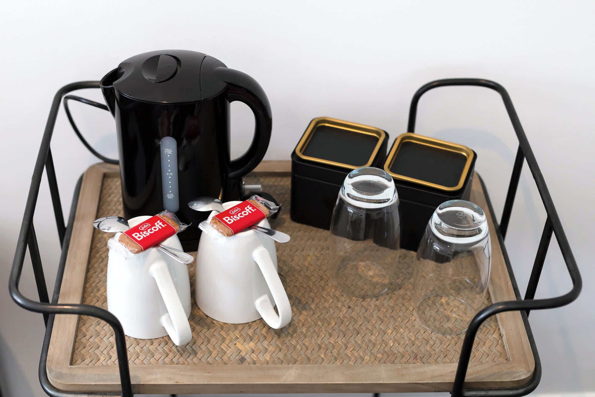 Una bandeja con una cafetera, dos sobres de azúcar, dos tazas blancas, dos frascos de vidrio y dos recipientes negros.