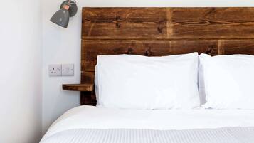 Una cama con ropa blanca, cabecera de madera, lámpara de noche y un enchufe en la pared.
