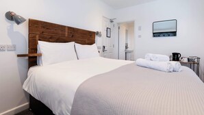 A hotel room with a wooden headboard, a large bed with white linens, a bedside table with a lamp, a mirror, and a small kitchenette area with a coffee maker and towels. - The Nook - Room 4 - Deluxe King Room (St Ives)