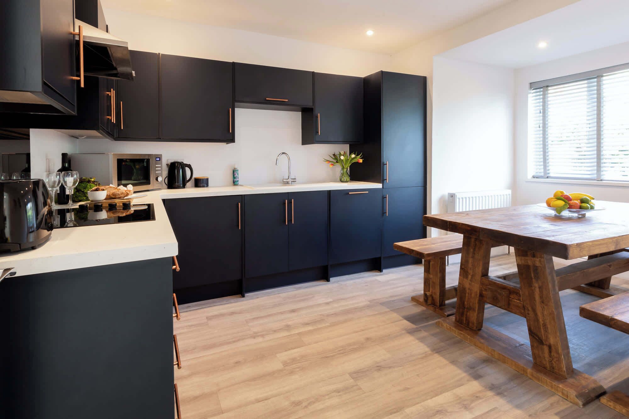A modern kitchen with dark cabinets, a wooden dining table, and a window with blinds.