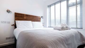 A hotel room with a wooden headboard, white bedding, a window with blinds, and a rolled towel on the bed.