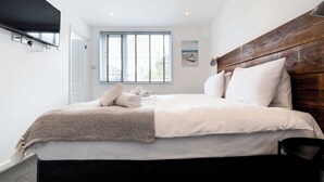 A modern hotel room with a large bed, a wooden headboard, a flat-screen TV mounted on the wall, and a window with blinds.