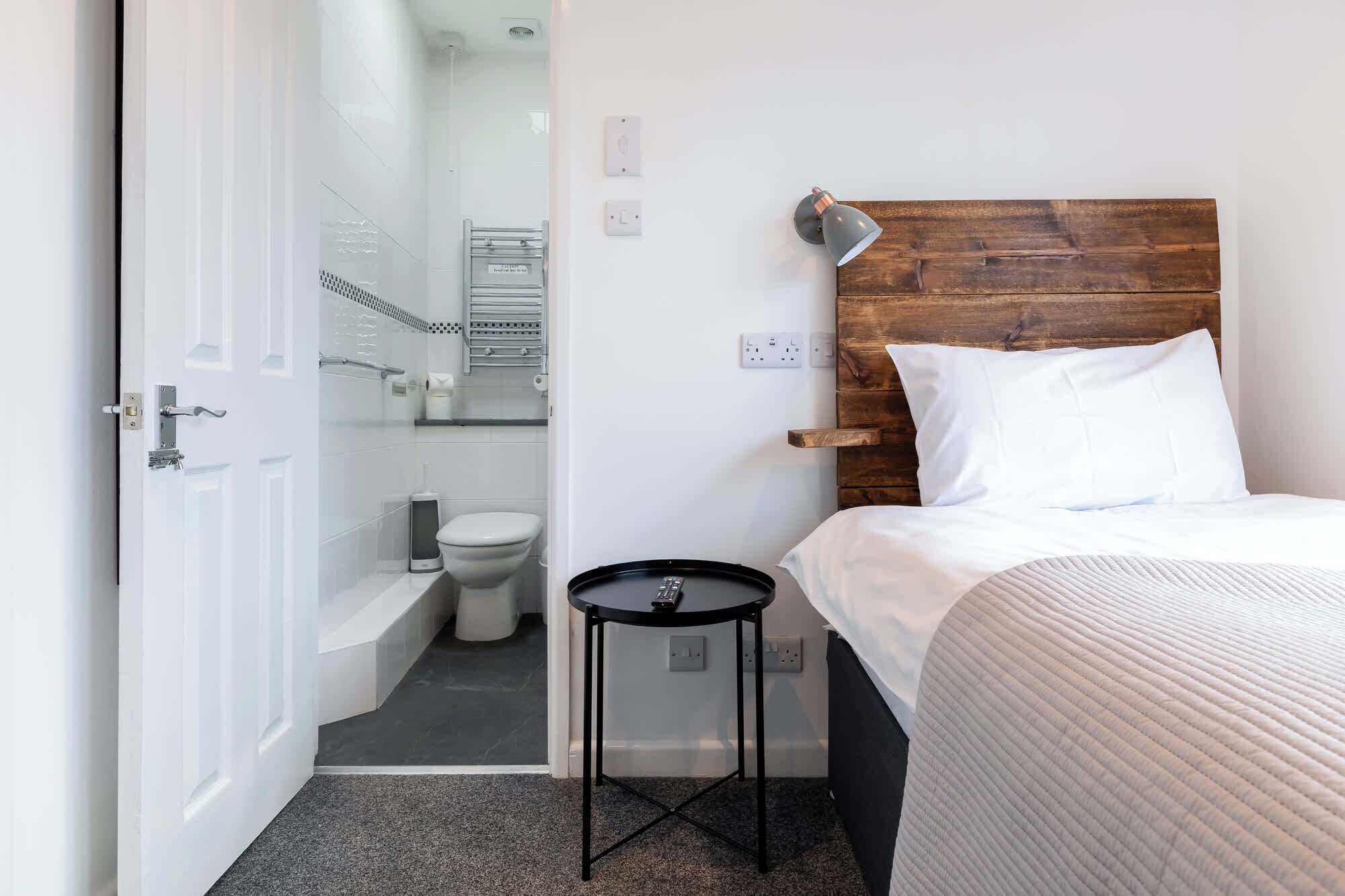 A room with a bed, a small table, a wooden headboard, a bathroom with a toilet and shower, and a door.