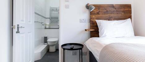 A room with a bed, a small table, a wooden headboard, a bathroom with a toilet and shower, and a door.