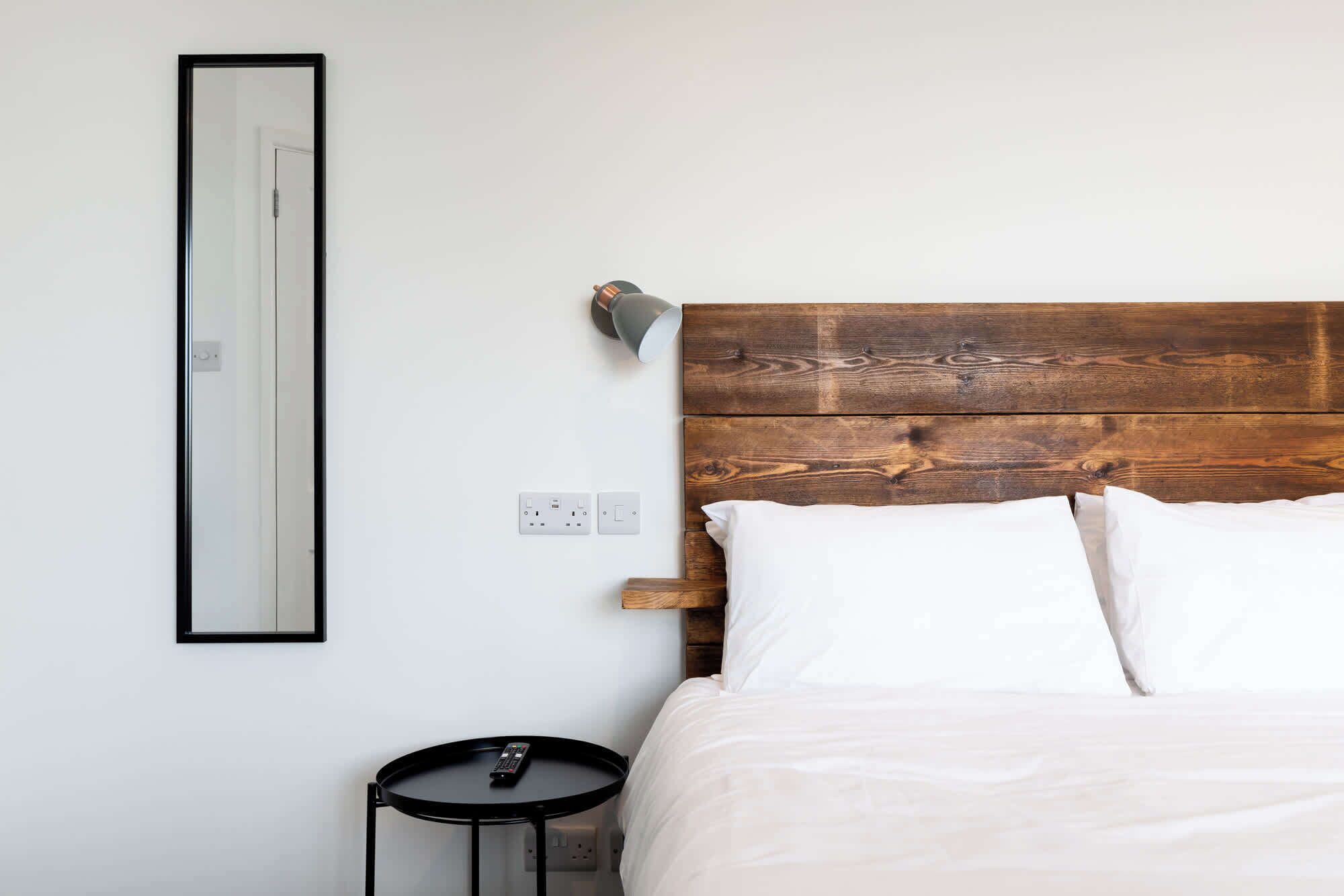 A bedroom with a wooden headboard, white bedding, a round black table, and a wall-mounted light fixture.