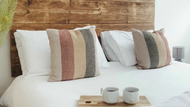 Ein ordentlich bezogenes Bett mit Holz Kopfteil, zwei Kissen, einem Tablett mit zwei Tassen und einem gerollten Handtuch.