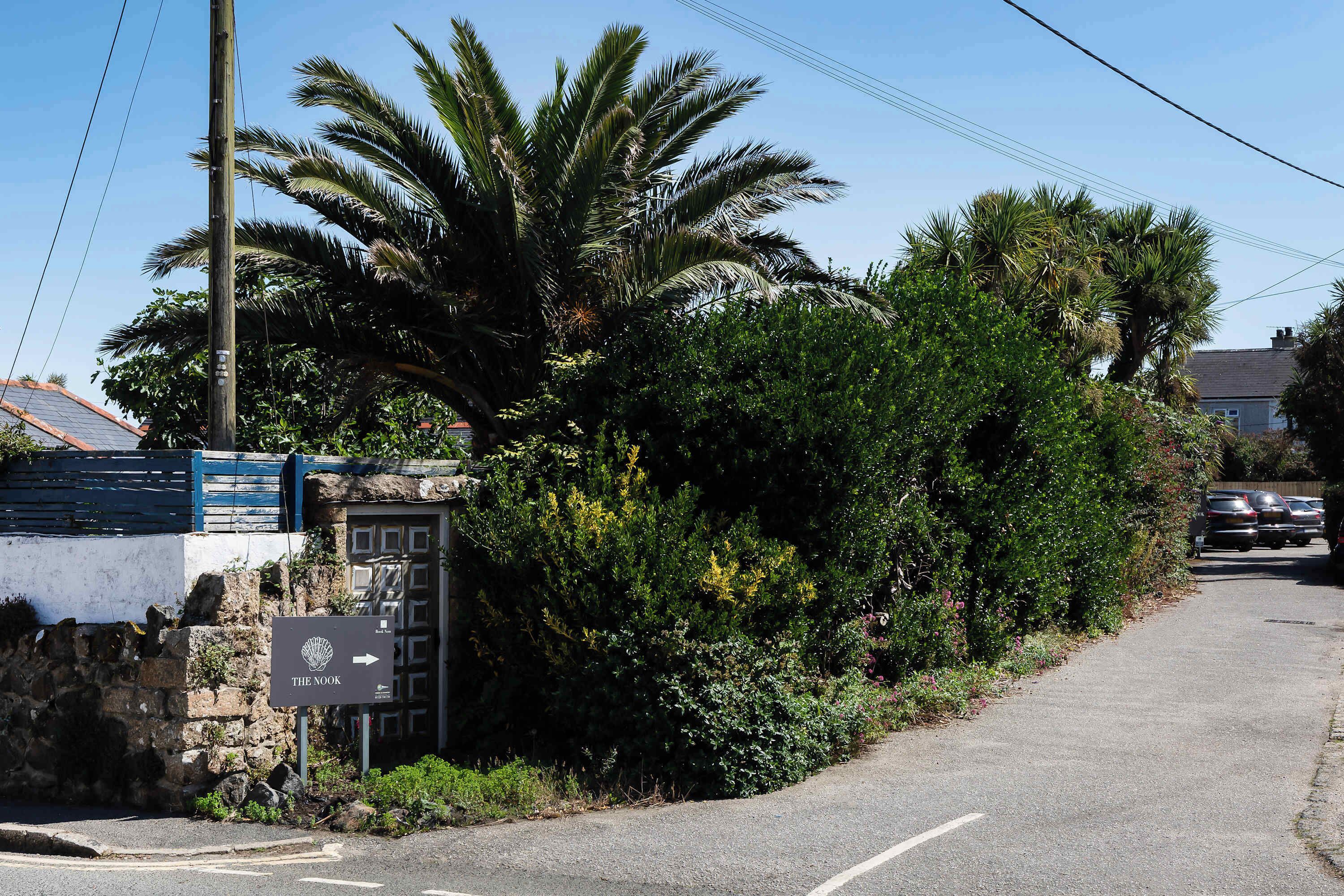 A street with a sign for 'The Nook,' a stone wall, and lush greenery including palm trees.