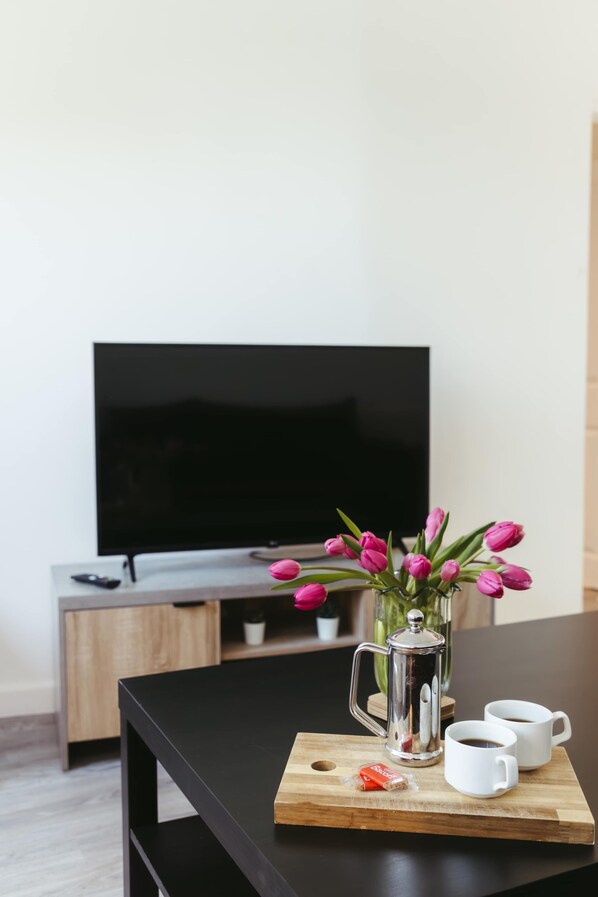 Una mesa de madera con una cafetera, dos tazas blancas y tulipanes rosados. En el fondo, un televisor está montado sobre un mueble de madera.