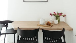 Ein schlichter Essbereich mit Holztisch, schwarzen Stühlen, einem gerahmten Bild und einer Vase mit Blumen.