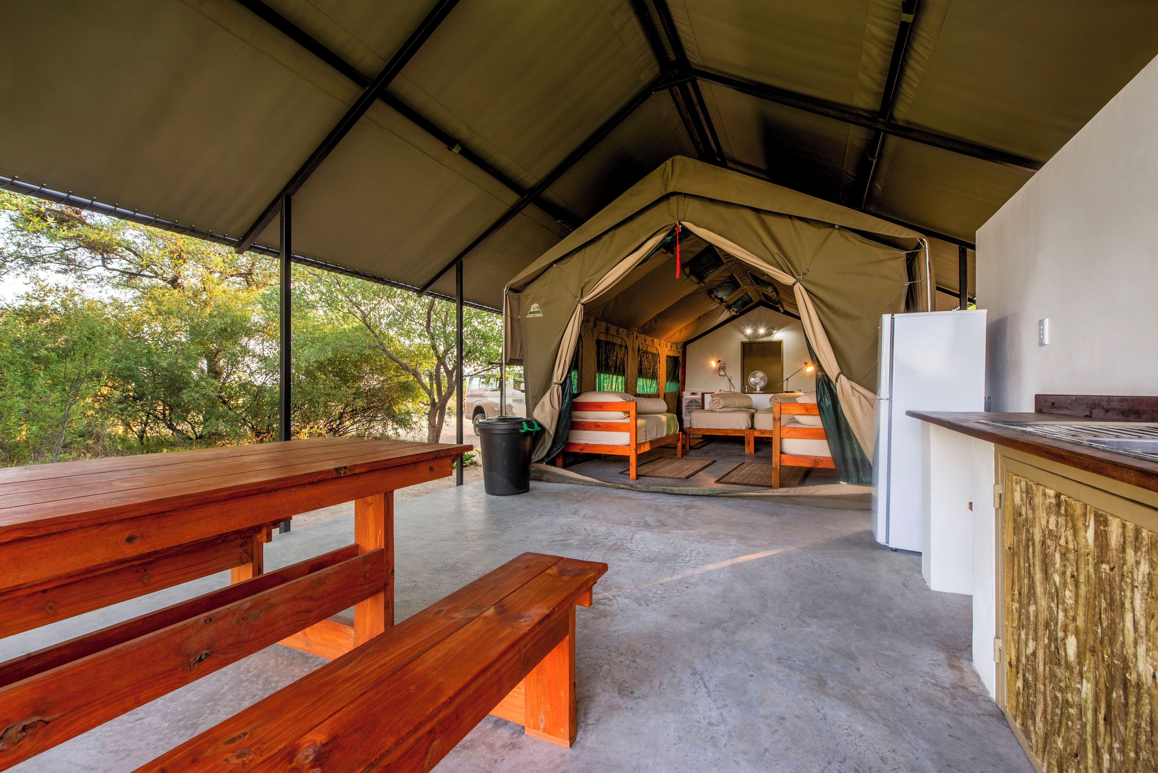 A tented accommodation with a wooden bench, a refrigerator, and a window overlooking greenery.