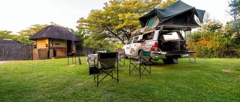 A vehicle with a tent and camping gear, a thatched-roof structure, and a grassy yard.
