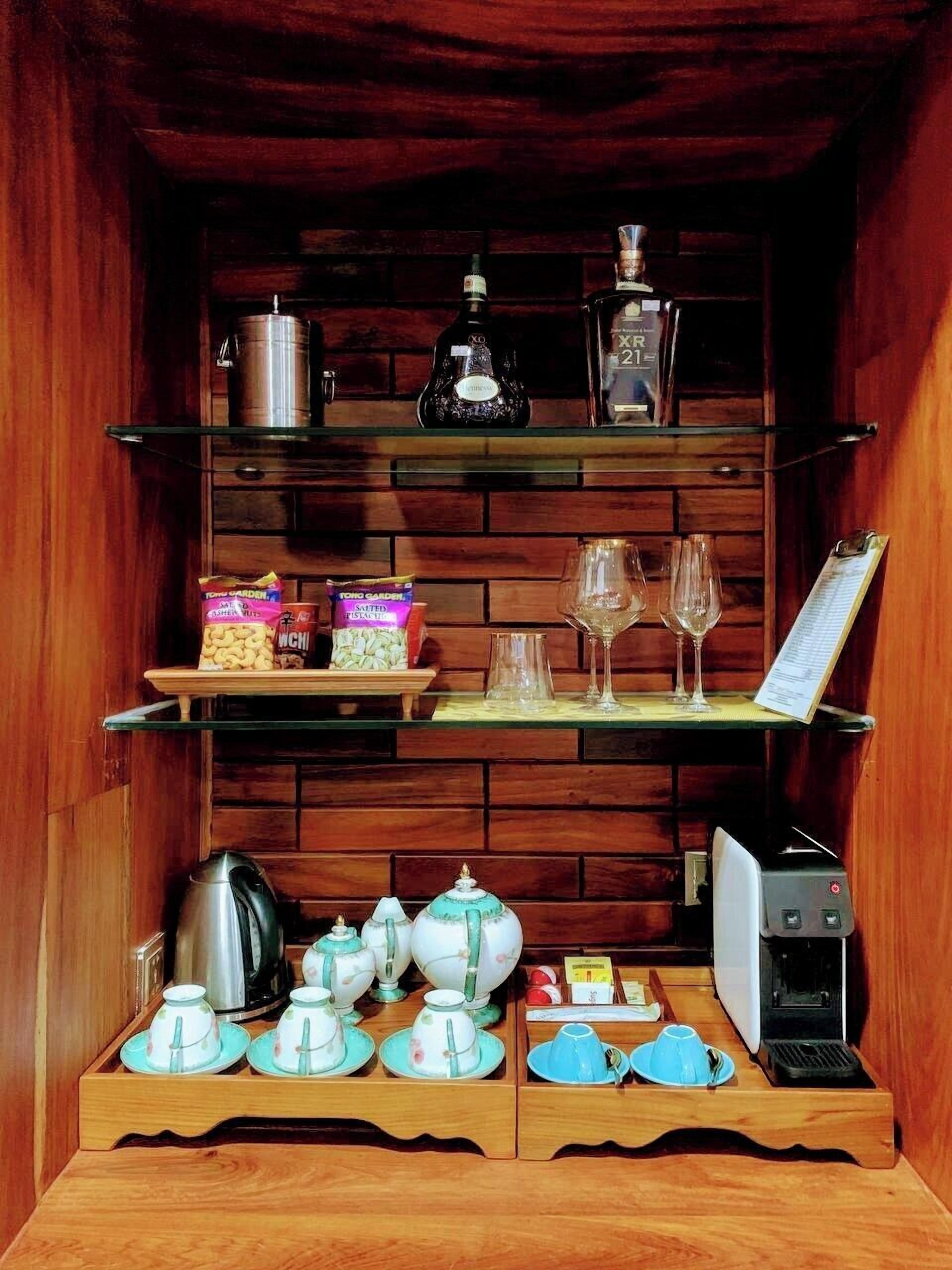 A compact kitchen with wooden cabinetry, a coffee machine, tea sets, and a shelf with glassware and snacks.