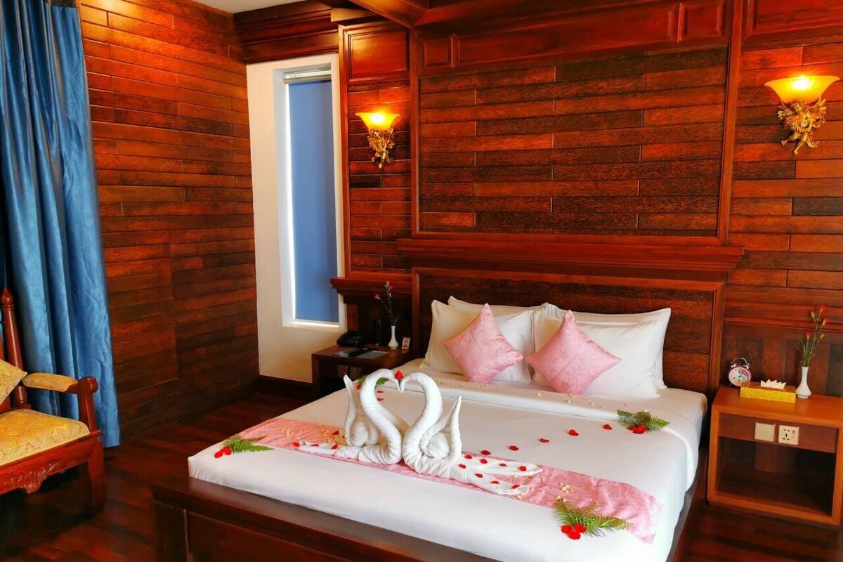 Een hotelkamer met een houten wandpaneel, een bed met witte lakens en roze kussens, een nachtkastje met een lamp en een stoel met een geel kussen.