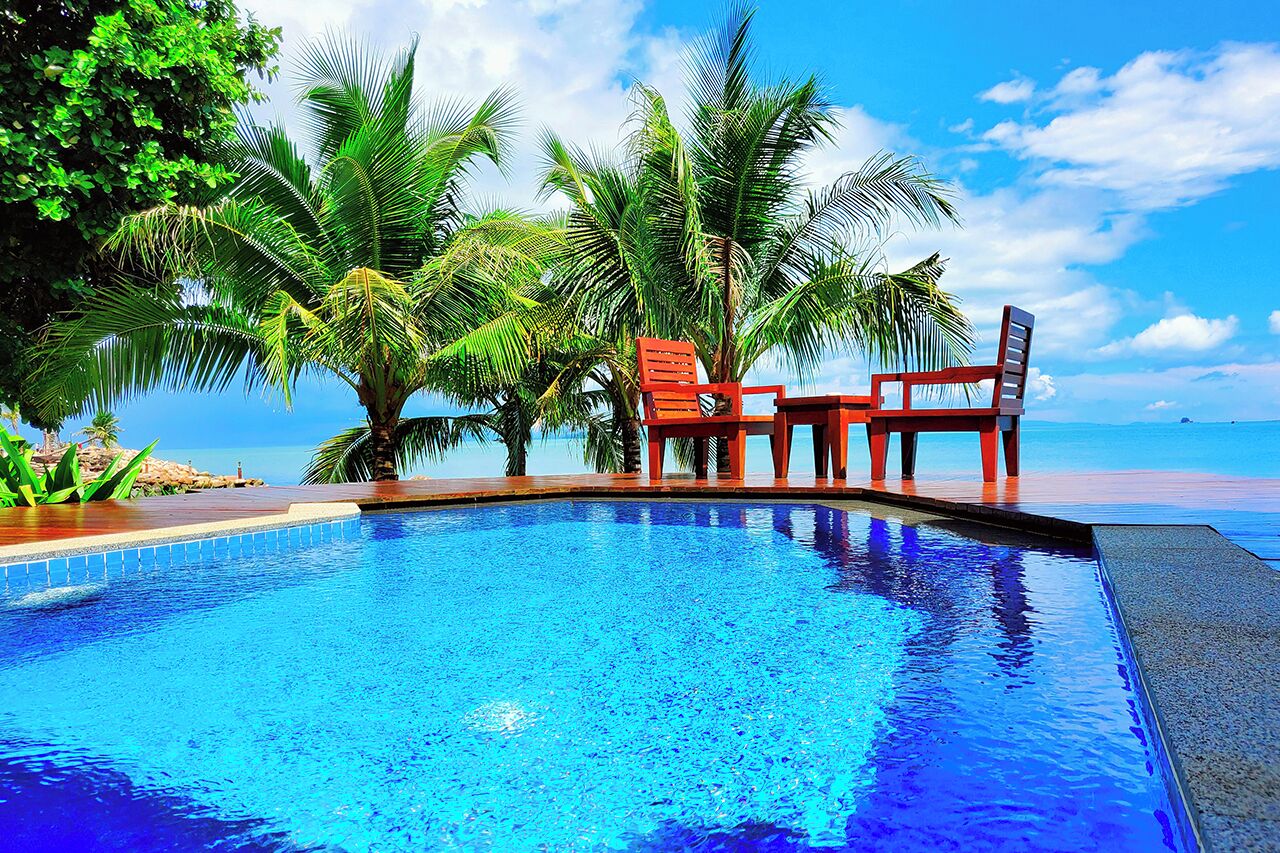 A swimming pool with two red chairs and a table, surrounded by palm trees, with a clear view of the ocean.