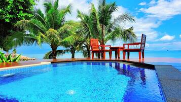 A swimming pool with two red chairs and a table, surrounded by palm trees, with a clear view of the ocean.