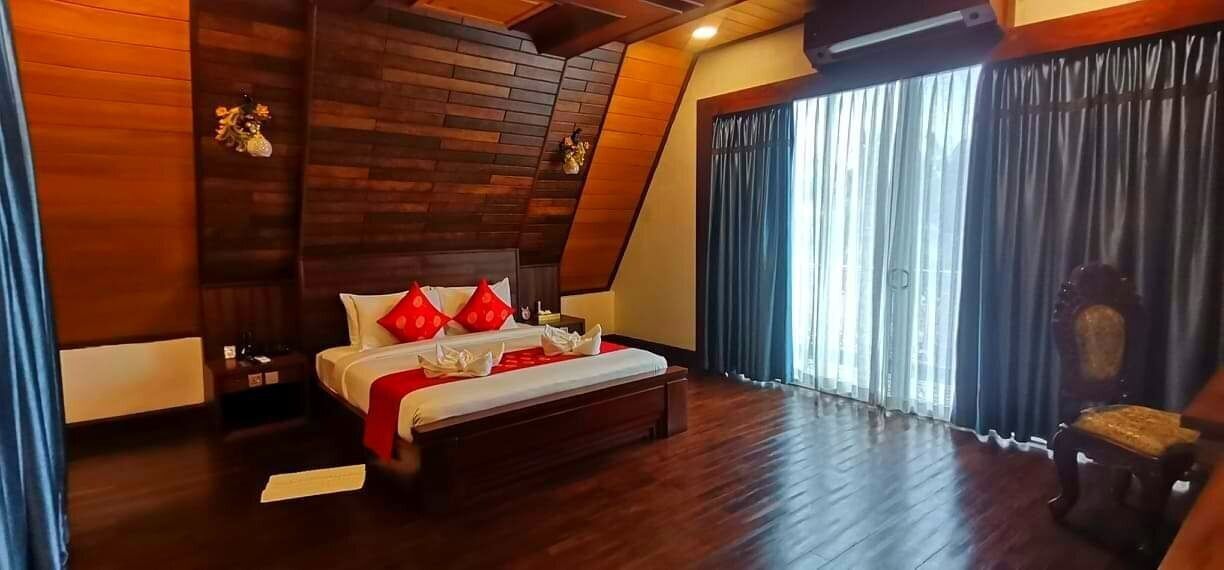 A bedroom with a wooden ceiling, a large bed with red pillows, and a view of the outdoors through a window with curtains.
