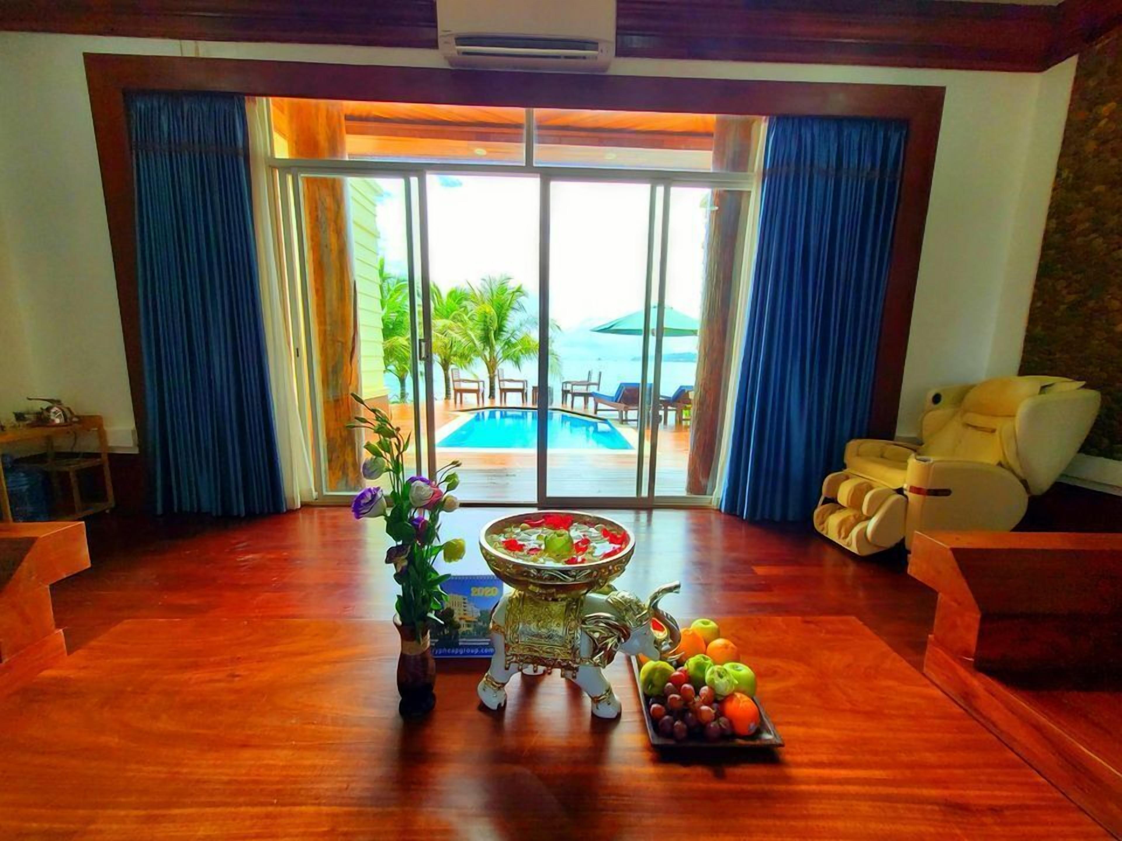 A spacious room with a wooden floor, a large window with blue curtains, a pool view, a massage chair, and a fruit bowl on a pedestal.