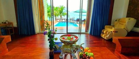 A spacious room with a wooden floor, a large window with blue curtains, a pool view, a massage chair, and a fruit bowl on a pedestal.