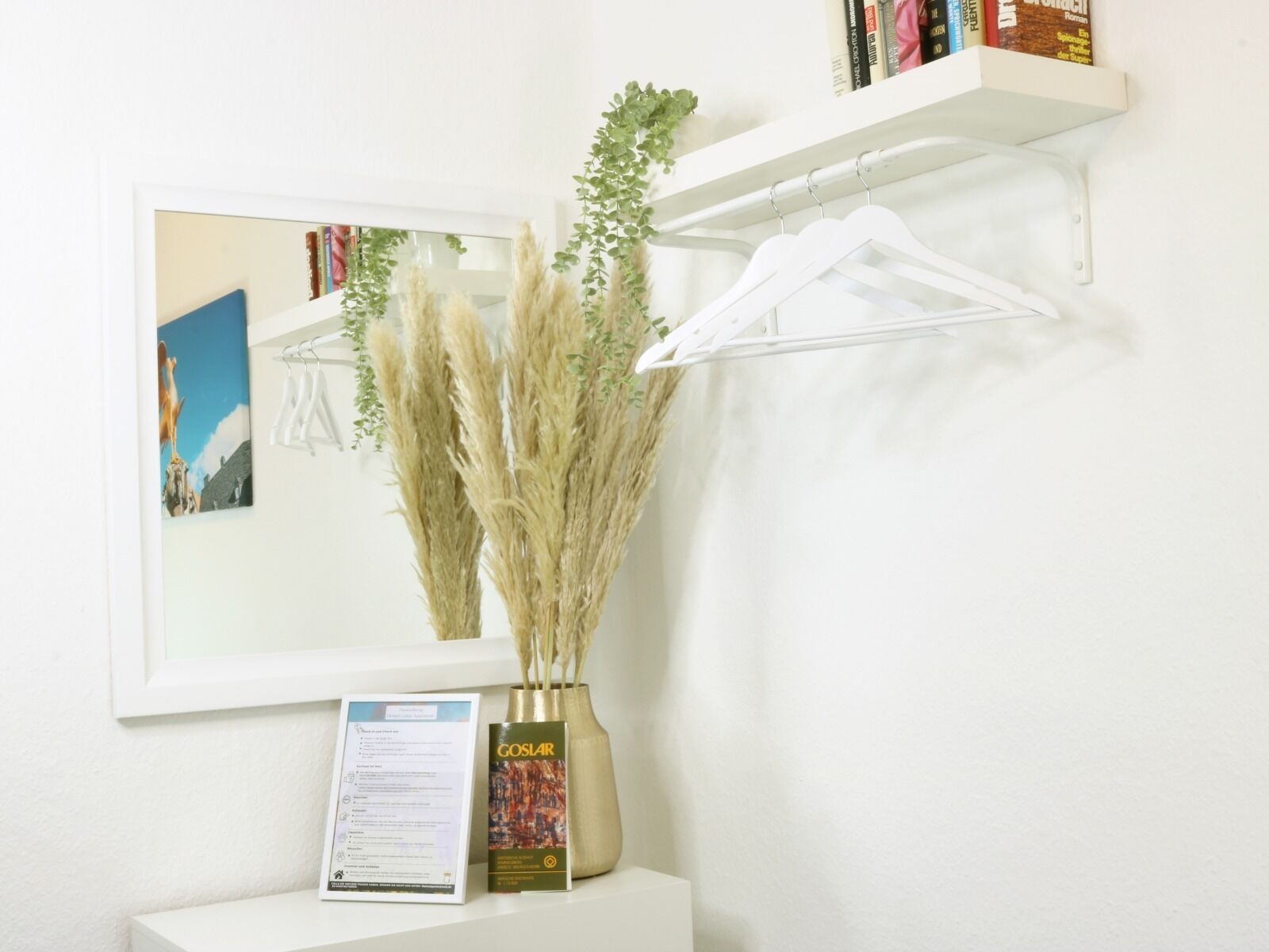 Un mur blanc orné d’un tableau encadré, une plante suspendue, une étagère blanche avec des livres et un tableau blanc.