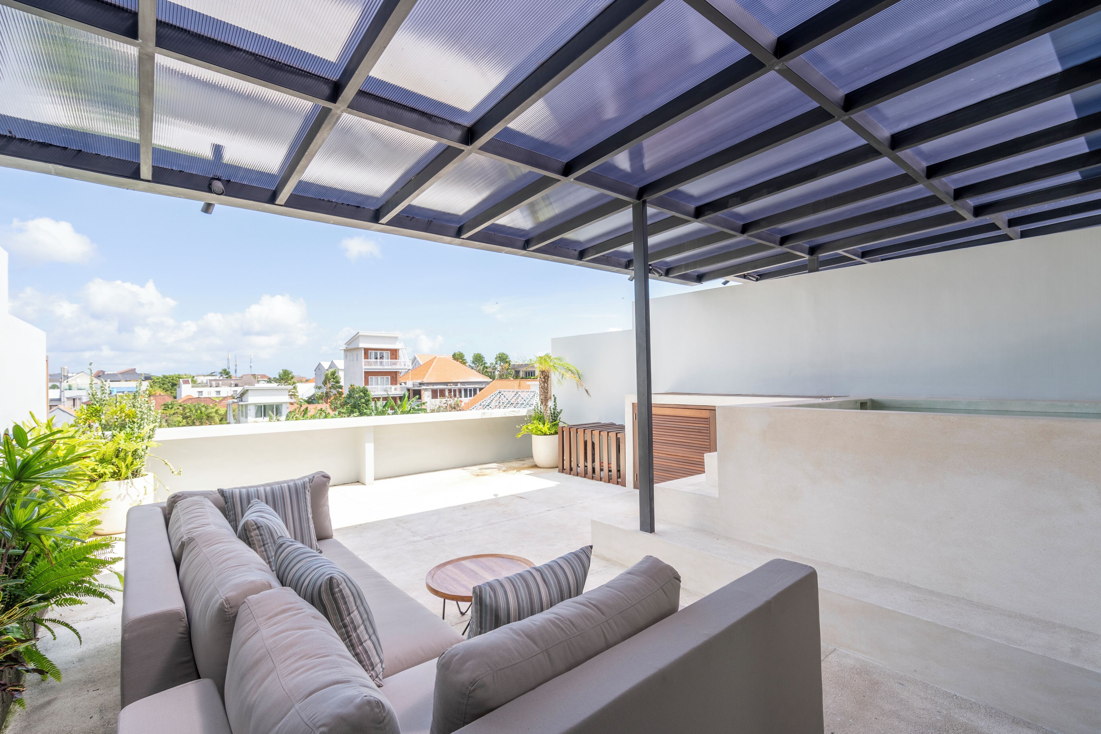 Ein moderner Außenbereich mit Sitzecke, einer Couch und einem kleinen Tisch, einer Pergola darüber und Blick auf Gebäude in der Ferne.
