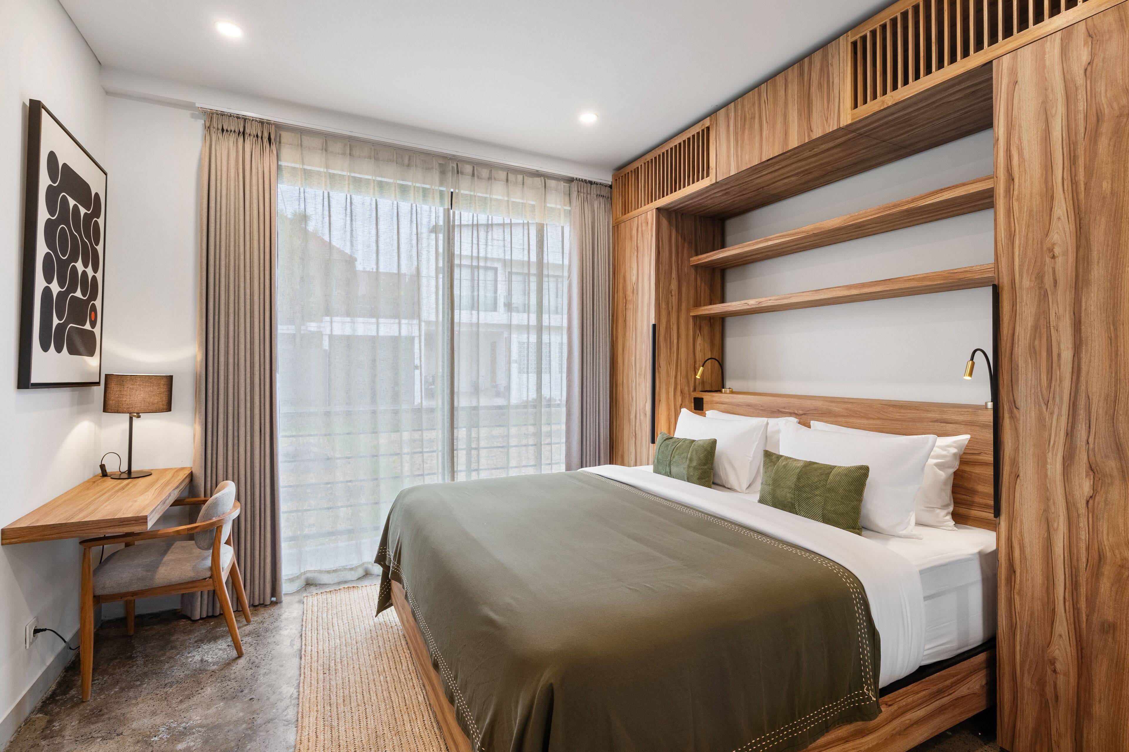 A hotel room with a wooden headboard, a desk with a chair, a bed with white and green pillows, and a large window with sheer curtains.